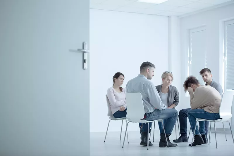 Image: stark stock photo with much white space showing a woman crying in a small seated group at a rehab center with an open door in the foreground
